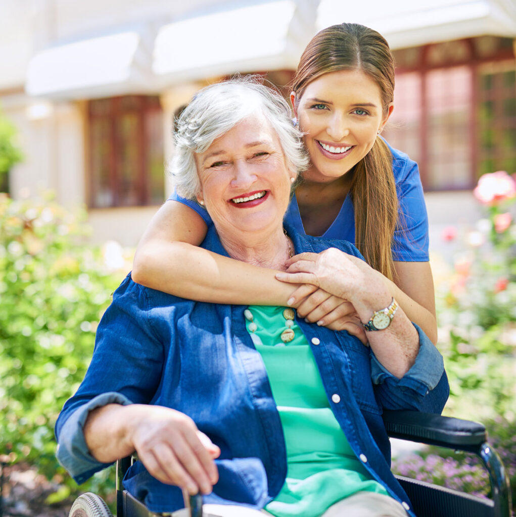 Nurse wearing blue scrubs hugging older woman wearing teal whirt under dark blue buttoned shirt sitting in a wheelchair outside