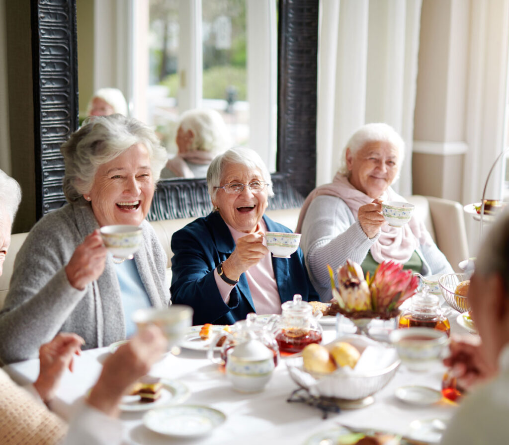 Three older adult ladies sitting at a table with food on it while holding teacups and smiling to others at the table not in view