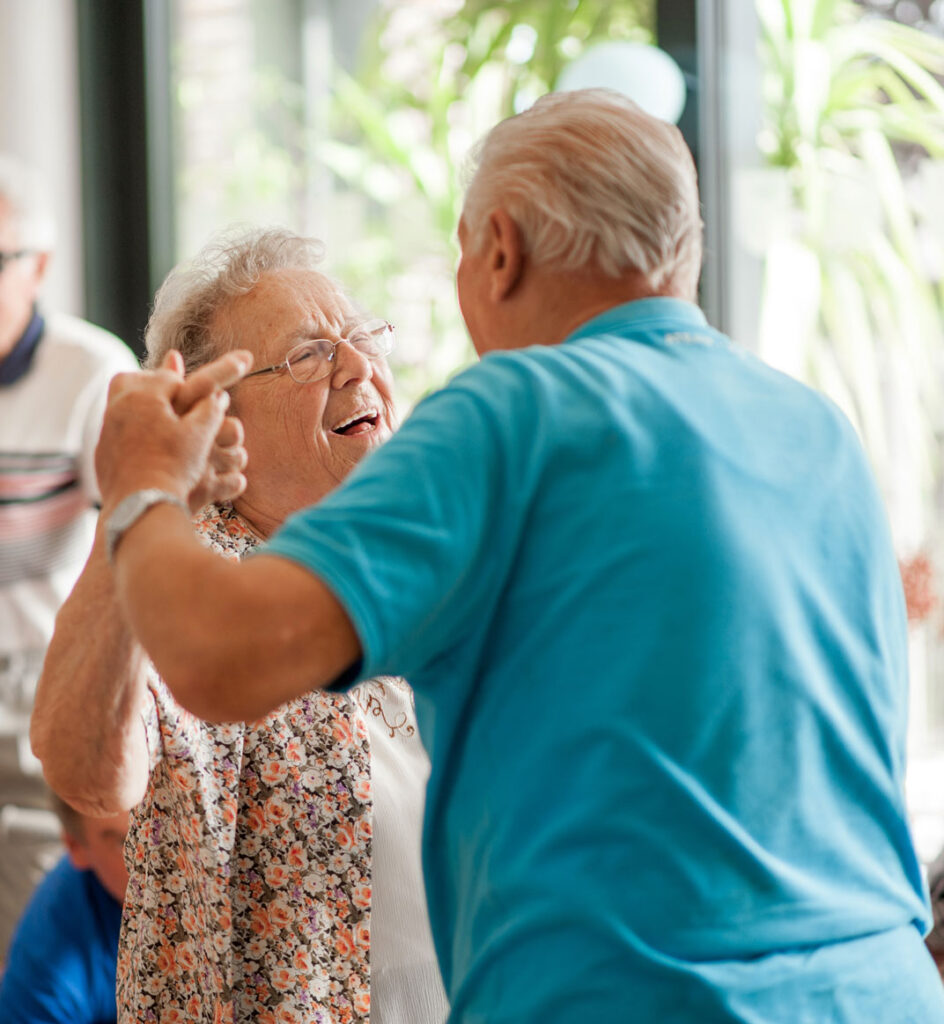 Older adult couple holding hands while staring and smiling at eachother dancing with man in glasses out of focus in background
