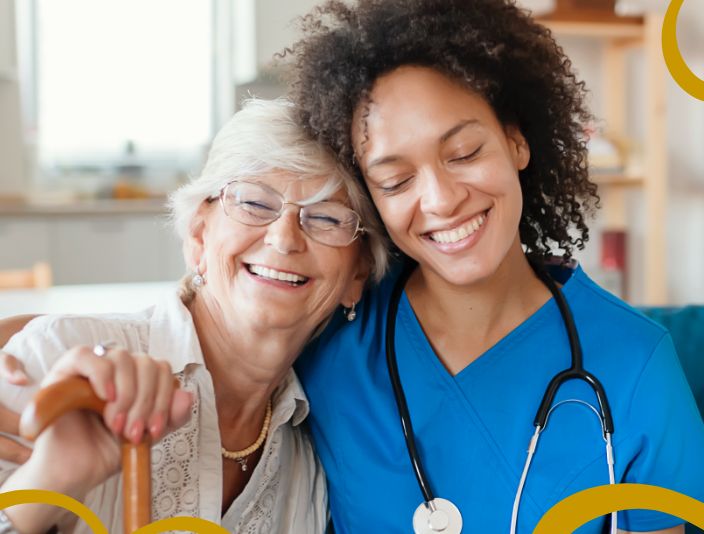 Nurse wearing blue scrubs and a stethescope smiling with resident who is leaning on her shoulder